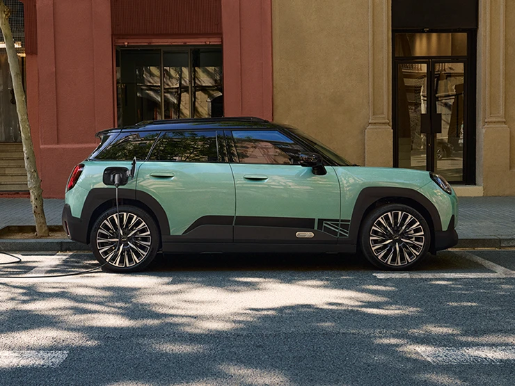 Страничен изглед на изцяло електрическото MINI Aceman в цвят Ocean Wave Green с черен покрив, което зарежда на улицата.