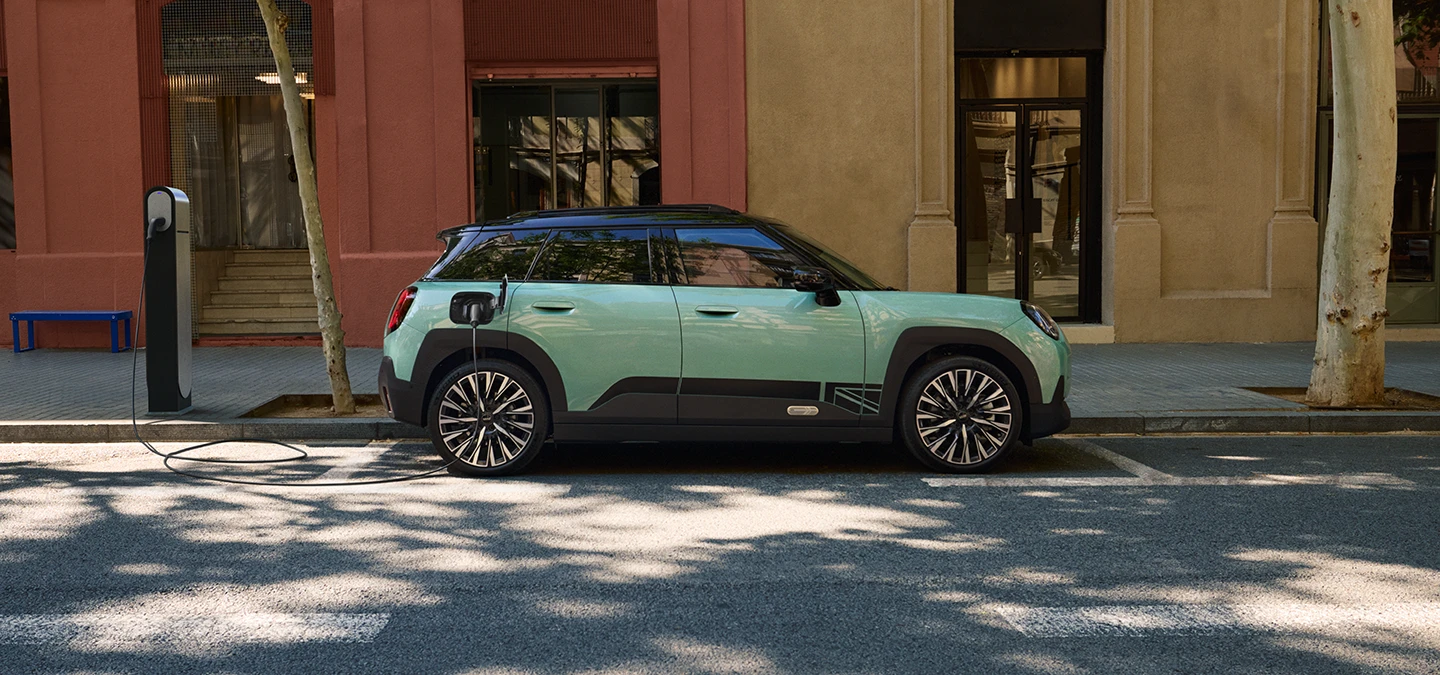 Страничен изглед на изцяло електрическото MINI Aceman в цвят Ocean Wave Green с черен покрив, което зарежда на улицата.