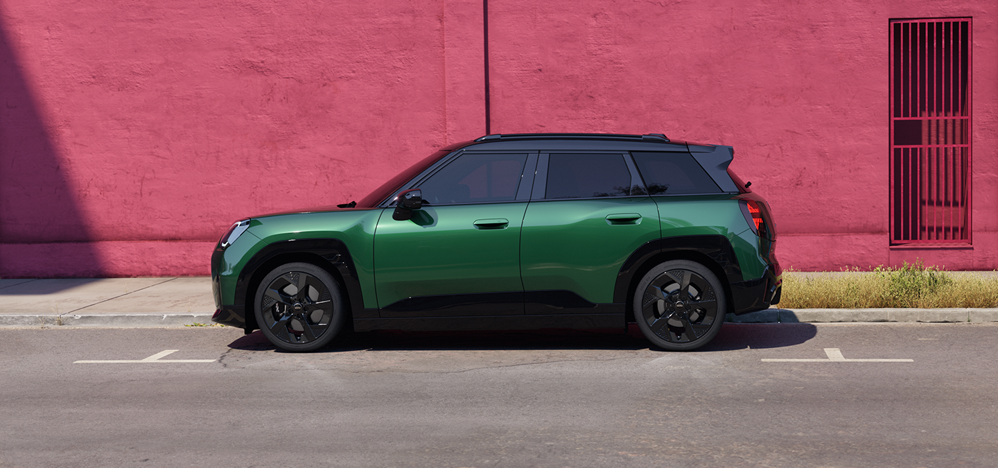 Страничен изглед на напълно електрическото MINI Aceman в цвят British Racing Green с черен покрив на улица.