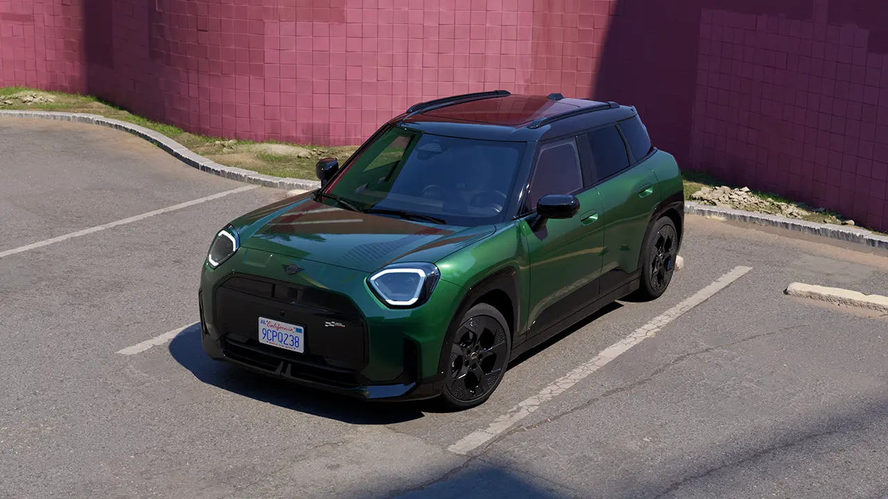 Изглед отпред на паркирано изцяло електрическо MINI Aceman в цвят British Racing Green на паркинг с розова стена на заден план