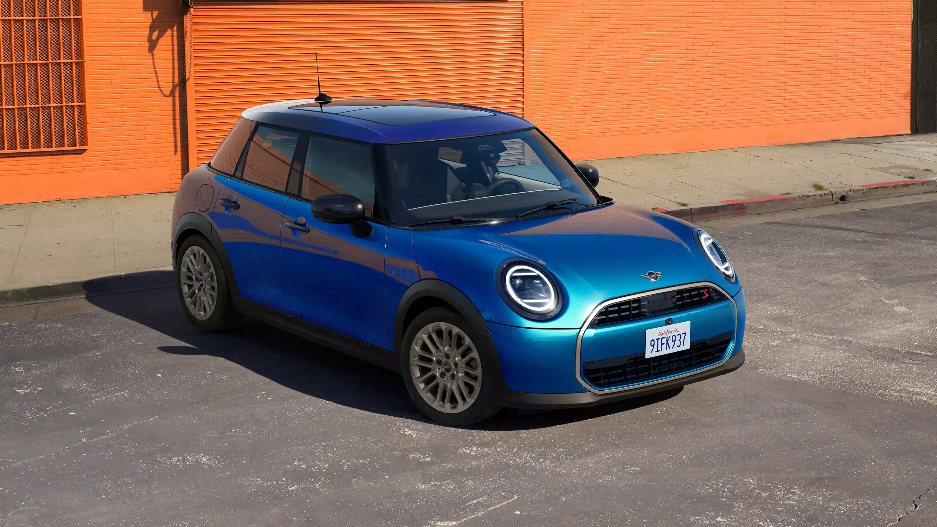 Изглед отпред на паркиращо MINI Cooper 5-Door в цвят Icy Sunshine Blue с покрив Multitone Blue на улицата с оранжева стена на фон