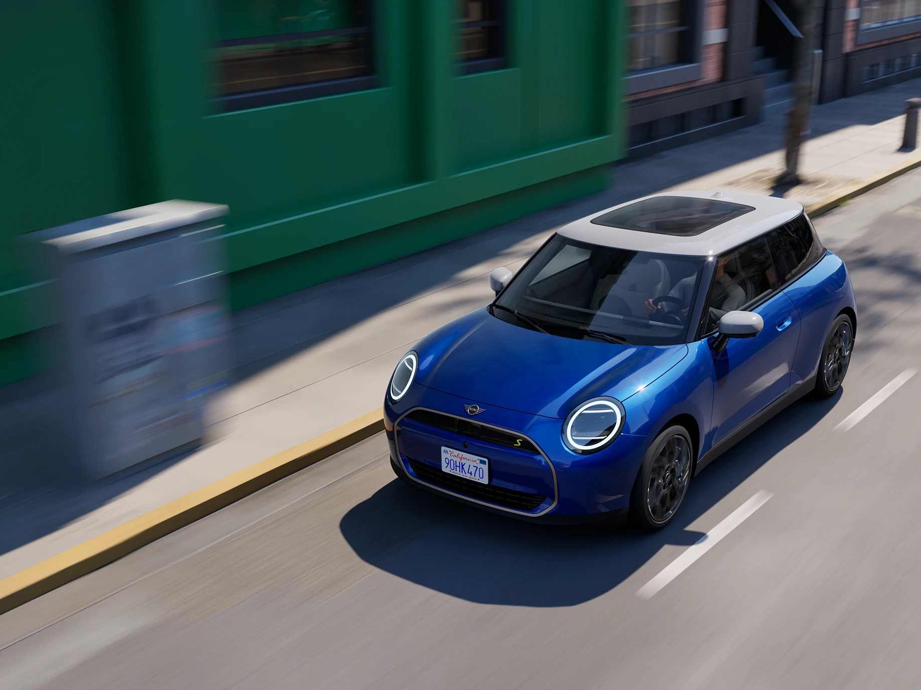 Изглед отгоре на изцяло електрическото MINI Cooper в цвят Blazing Blue с бял покрив, движещо се по улица. 