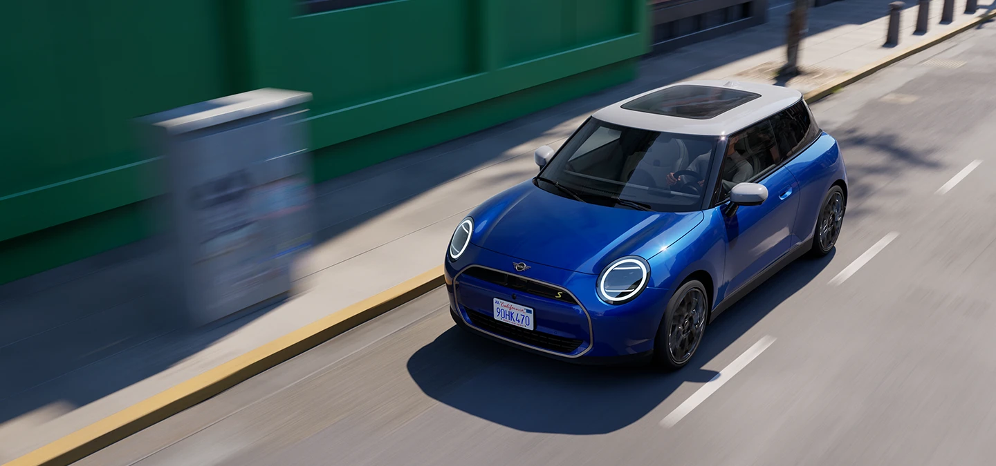 Изглед отгоре на изцяло електрическото MINI Cooper в цвят Blazing Blue с бял покрив, движещо се по улица. 
