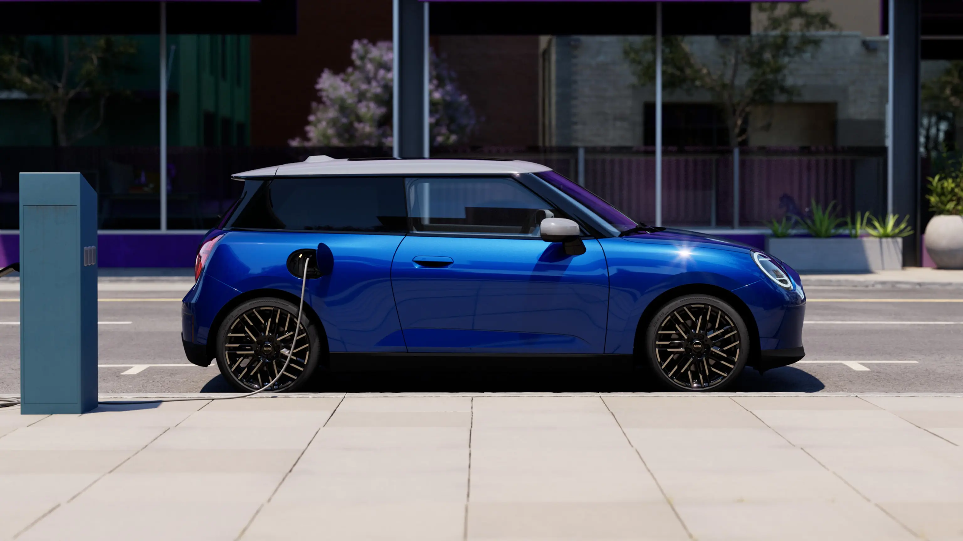 Страничен изглед на напълно електрическо MINI Cooper в цвят Blazing Blue, паркирано на тротоар до зарядна станция
