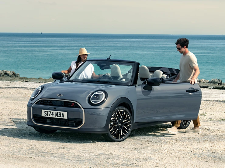 Изглед отпред вляво на MINI Cooper Cabrio в цвят Copper Grey на плажа с двама души до него.