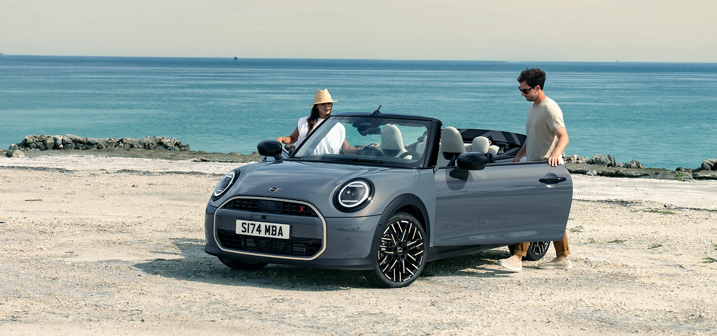 Изглед отпред вляво на MINI Cooper Cabrio в цвят Copper Grey на плажа с двама души до него.