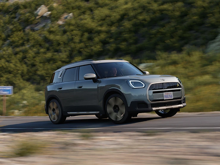 Страничен изглед на напълно електрическото MINI Countryman в цвят Smokey Green с покрив в цвят Vibrant Silver, което се движи извън пътя.