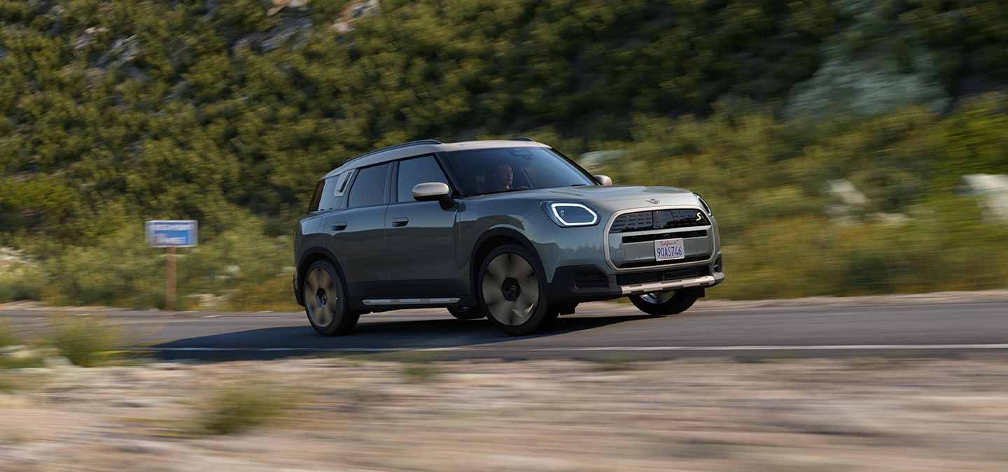 Страничен изглед на напълно електрическото MINI Countryman в цвят Smokey Green с покрив в цвят Vibrant Silver, което се движи извън пътя.