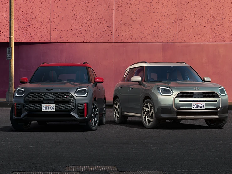 Изглед отпред на MINI Countryman JCW в Legend Grey с червен покрив Legend и MINI Countryman в цвят Smokey Green с покрив в цвят Vibrant Silver, стоящи едно до друго.