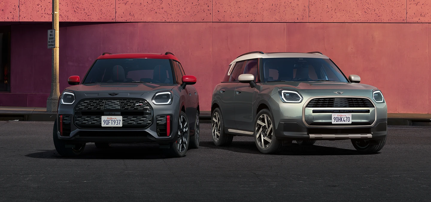 Изглед отпред на MINI Countryman JCW в Legend Grey с червен покрив Legend и MINI Countryman в цвят Smokey Green с покрив в цвят Vibrant Silver, стоящи едно до друго.