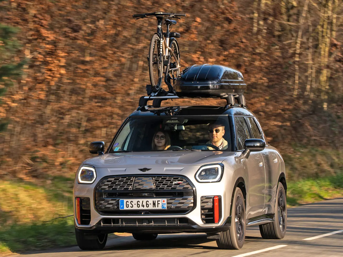 MINI Countryman Cooper в цвят Silver на планински път с багажник за покрив MINI, кутия за багаж на покрива MINI, багажник за велосипеди на покрива MINI с монтиран един велосипед плюс MINI John Cooper Works winglet – изглед отпред. 
