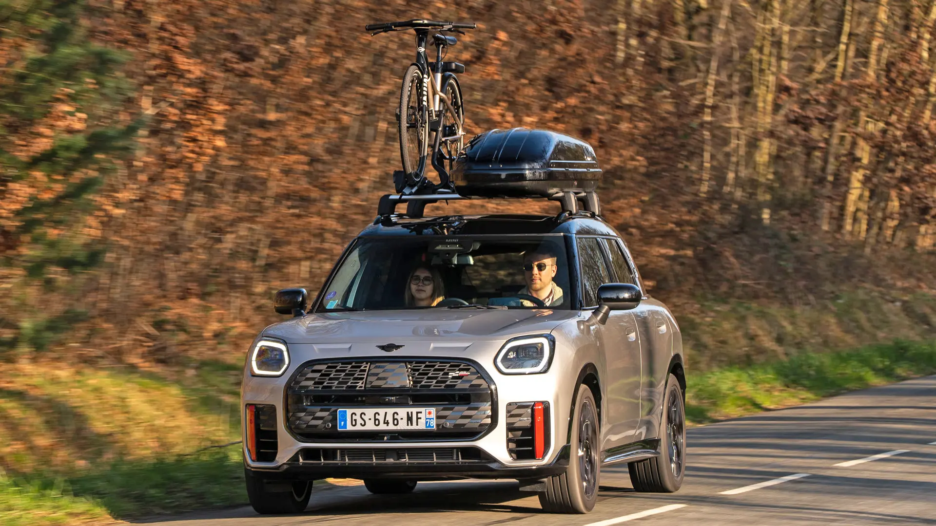 MINI Countryman Cooper в цвят Silver на планински път с багажник за покрив MINI, кутия за багаж на покрива MINI, багажник за велосипеди на покрива MINI с монтиран един велосипед плюс MINI John Cooper Works winglet – изглед отпред. 