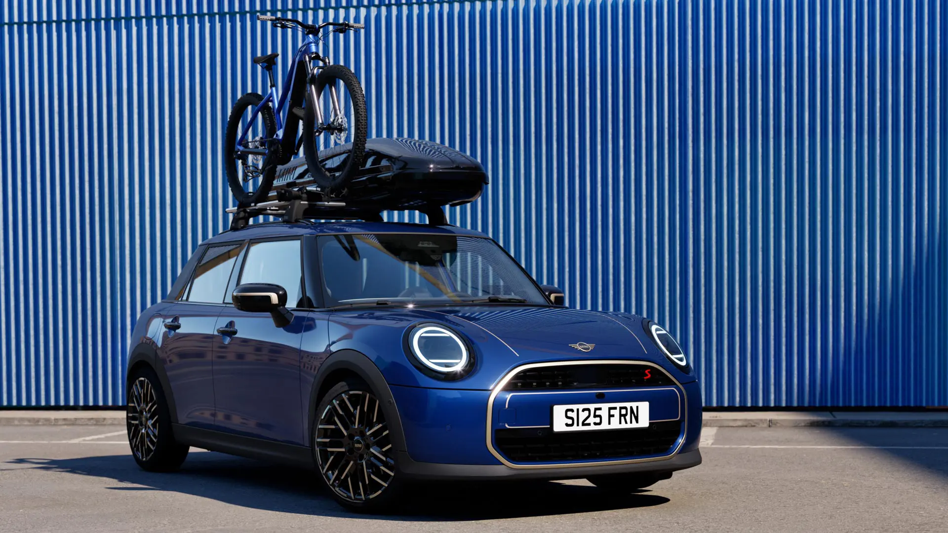 Изглед отпред на MINI Aceman в цвят Indigo Sunset Blue с багажник за велосипеди за покрива MINI, на който е закрепен планински велосипед, и кутия за багаж на покрива MINI 320.