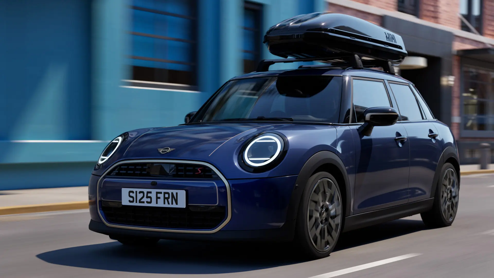 Три четвърти изглед отпред на MINI Aceman в цвят Indigo Sunset Blue, оборудвано с MINI багажник за покрива, на който е монтирана кутия за багаж на покрива MINI 320. 