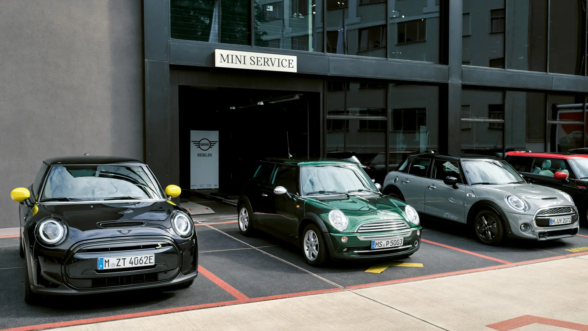 MINI Cooper Electric S в черен цвят пред център на MINI заедно с MINI Cooper с 3 врати в зелен цвят, MINI с 5 врати в сребрист цвят, MINI JCW в черен цвят, изглед отпред. 