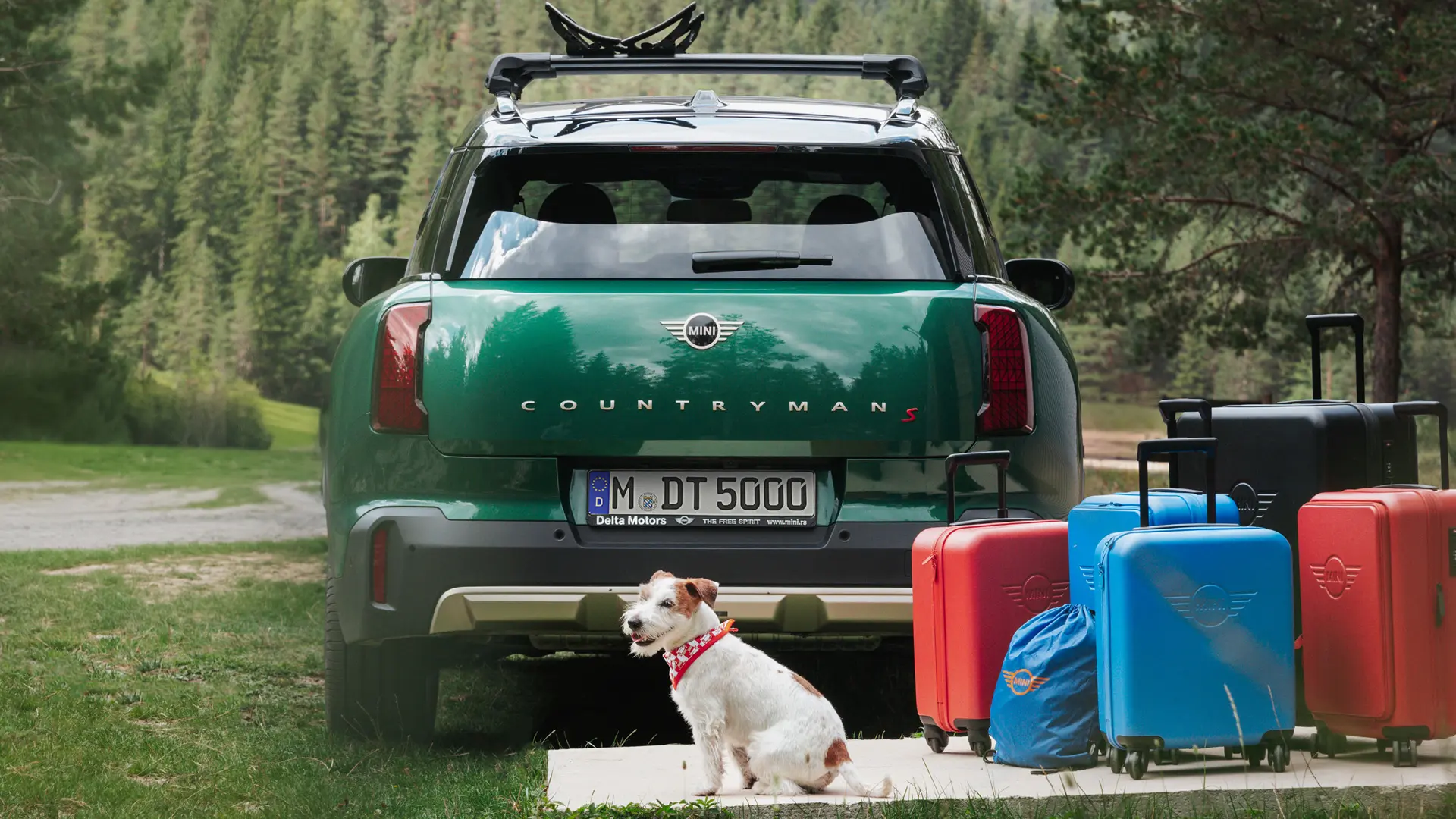 MINI Countryman в зелен цвят с разнообразни артикули за багаж от магазина на MINI Лайфстайл зад автомобила заедно с Джак Ръсел териер. 
