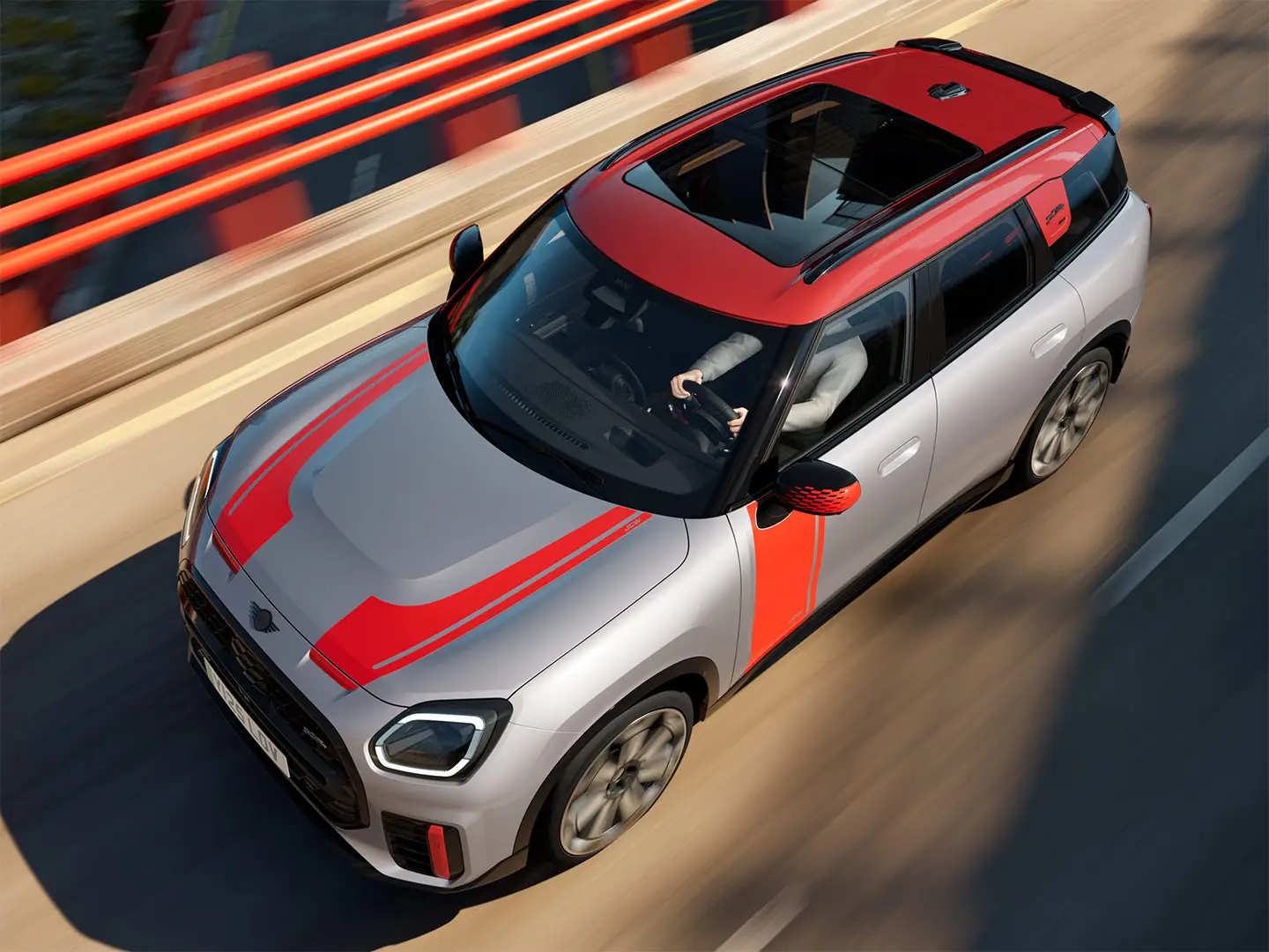 MINI Countryman John Cooper Works в цвят Melting Silver с капак на външното огледало и стикери отстрани и на капака, покрив и Winglet в цвят Chilli Red, което се движи по градска улица – изглед отпред.